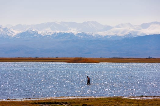 Alone fisherman checking nets in shallow waters of Issyk-Kul lake with mountains on background at autumn afternoon.