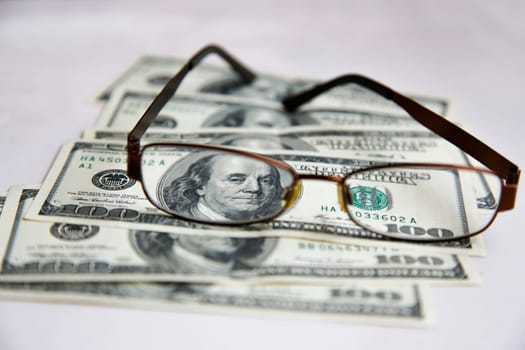 Close-up of eyeglasses resting on a stack of 100-dollar bills on a white background
