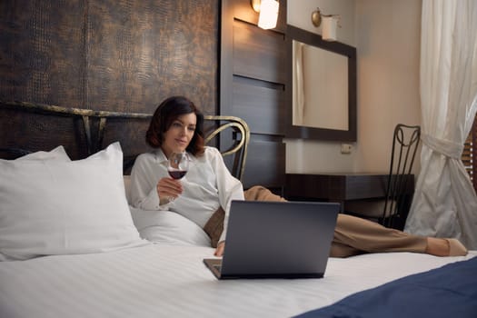 Relaxed businesswoman lying in bed drinking wine and browsing internet on laptop computer enjoying evening in hotel room