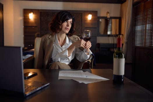Thoughtful businesswoman drinking wine in luxurious hotel room. Beautiful female entrepreneur looking aside thinking about future while resting indoors
