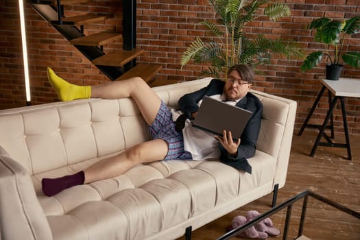 Surprised businessman grimacing looking at laptop screen working online sitting on sofa at home living-room. Confused salesman entrepreneur reading e-mail news using computer