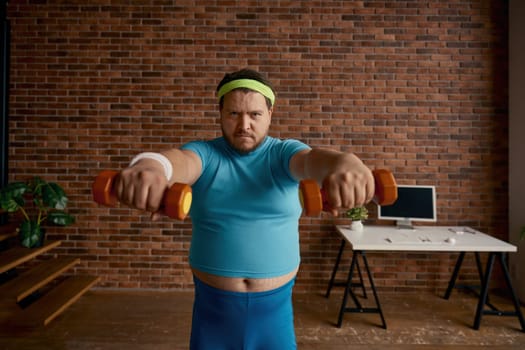 Portrait of serious comic man with obese figure doing workout with dumbbells at home. Cute fat sportsman healthy habits