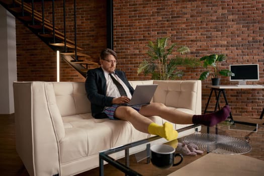 Portrait of funny businessman wearing formal suit and cute socks with different color working online on laptop. Concentrated salesman sitting on sofa using computer for internet job