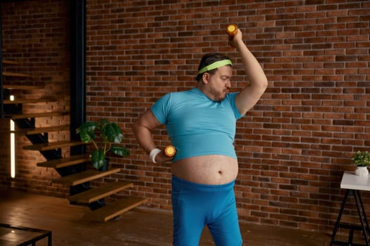 Fat man doing sport training with dumbbells at home. Funny hardworking guy exercising burning calories