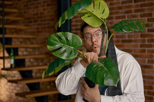 Portrait of funny businessman with necktie on head holding houseplant in hand. Stressful online work and alcohol abuse