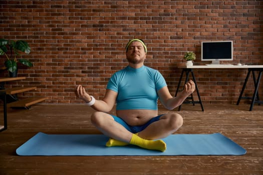 Relaxed fat sportsman meditating sitting on mat at home. Overweight man calming down practicing yoga in lotus position. Sports activity and weight loss