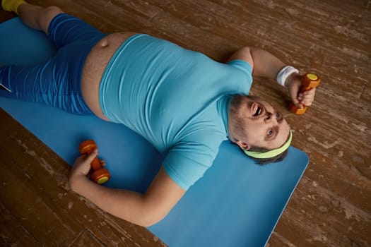 Funny overweight man feeling tired after slimming sports workout. Physical exercise with umbels for weight loss and muscle building