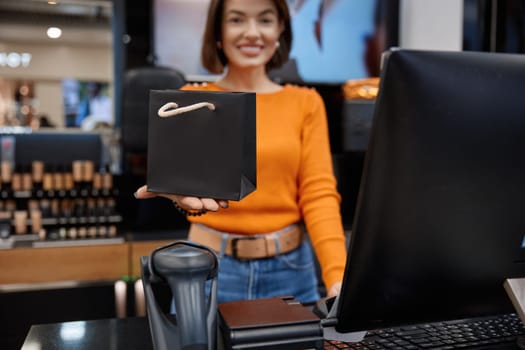 Smiling sales woman in cosmetics store holding out the package. Attractive positive young lady working with customers