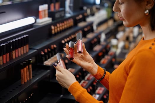 Attractive woman choosing lipstick in cosmetics store. Young lady looking for suitable decorative product for lips