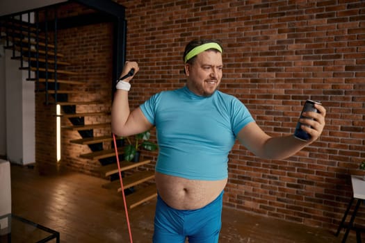 Funny overweight man training with fitness rubber drinking energy beverage. Gymnastics exercise at home