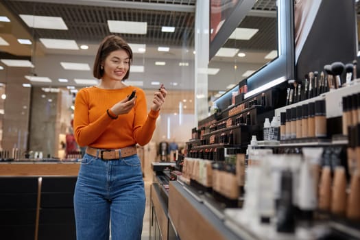 Positive woman choosing lipstick in cosmetics store. Young lady customer looking fore different decorative stuff for face