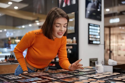 Smiling woman leaned to stand with eye shadow palettes different shades. Happy satisfied lady customer in cosmetics shop