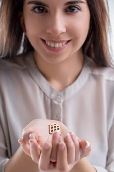 young woman with a bright smile holds a tiny house model in her palm. The image highlights real estate, home buying, love for ones home, insurance, and related financial matters, reflecting joy and