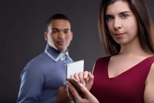 Young woman using her smartphone while standing in front of a blurred man using a tablet, both are on a gray background
