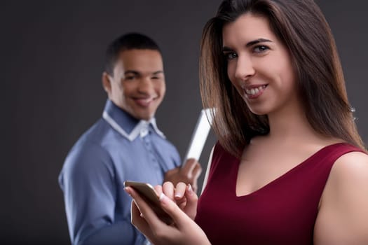 Two professionals, a businesswoman and businessman, smiling, using phone and tablet in office. Young adults show teamwork and success