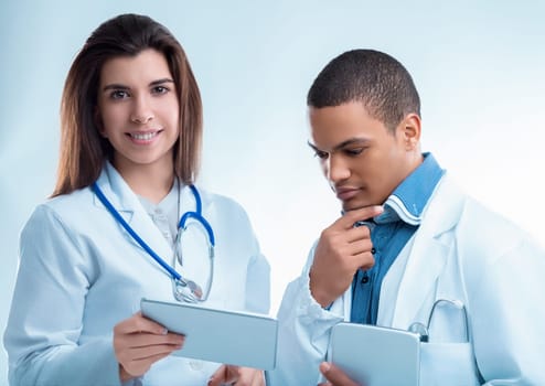 Two young doctors are using digital tablets while wearing white coats and stethoscopes. They are concentrating on the information on the screen