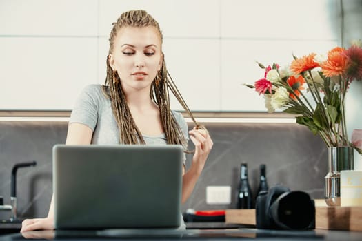 Young woman with long braids is working on a laptop in a modern kitchen, with her professional camera sitting on the counter beside her