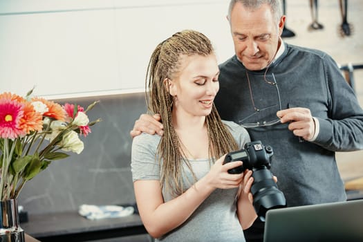 Photographer showing photos she took to a senior man, while holding a professional digital camera in a luminous kitchen