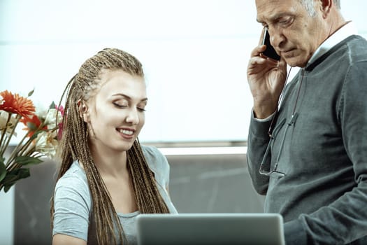 Young professional woman with long braided hair is concentrating on the laptop screen while her senior colleague is having a phone call