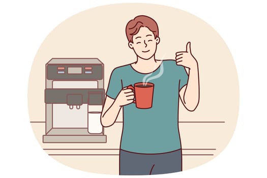 Man drinks coffee from mug standing near machine for making delicious espresso and shows thumbs up. Young guy takes coffee break to cheer up and increase productivity or stay awake doing tedious work