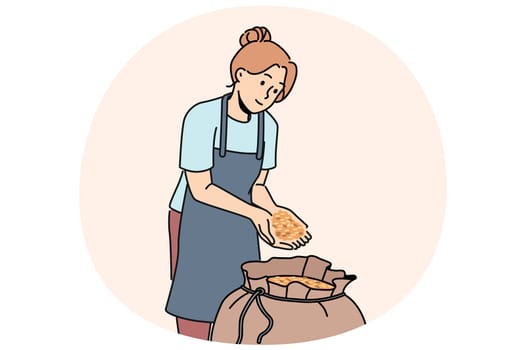 Woman stands near bag of malt for production of craft beer and collects hop grains in palms. Girl in apron works in agricultural factory preparing grains of prosso or wheat for processing.