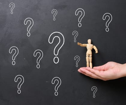 A woman's hand holding a wooden miniature puppet against a black chalkboard with chalk-drawn question marks. Concept of searching for truth, gathering information