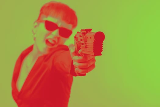 Woman with short hair and sunglasses is holding a gun, her mouth open in a scream, against a vibrant green background. The duotone effect adds a dramatic and artistic touch to the image