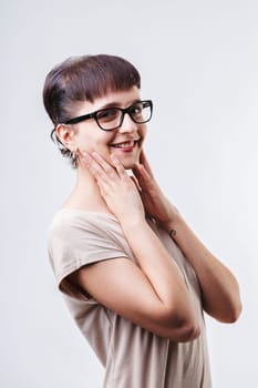 Young woman with short hair and glasses is touching her face and smiling on a white background. She is wearing a beige t shirt