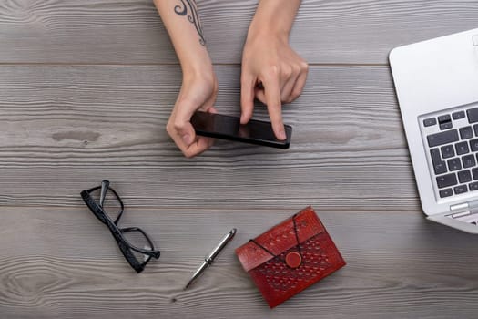 Hands holding a smartphone on a wooden desktop with a laptop, a notebook, a pen and eyeglasses, a flat lay image with copy space