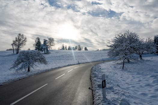 Snow landscape with trees, road and blue sky with clouds and sun