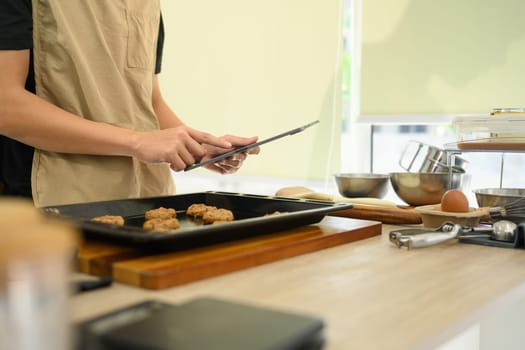 Young man preparing chocolate chip cookies in the kitchen and following recipe on digital tablet.