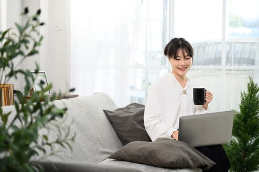 Cheerful asian woman sipping coffee and working on her laptop.