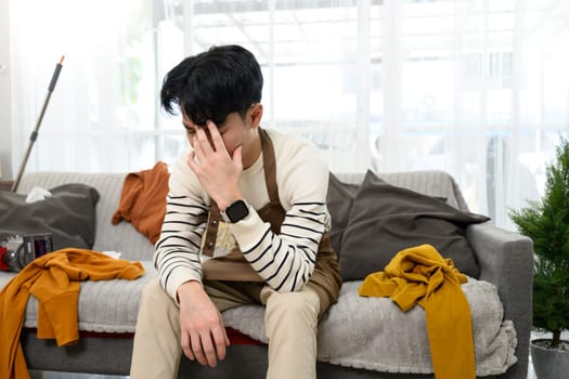 Tired and upset man sitting on couch in messy living room.