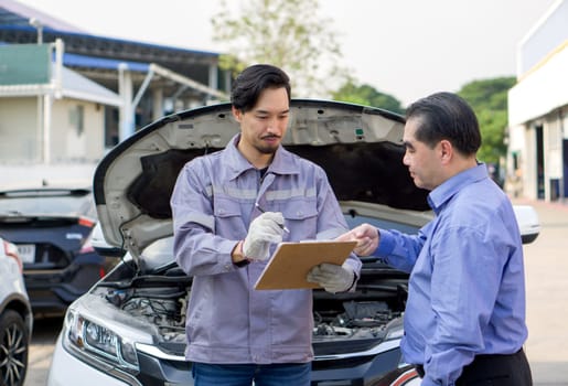 A mechanic in work uniform with gloves, holding document clipboard focused on the paperwork. The car owner or a service advisor discussing the vehicle's issues or maintenance work with the technician