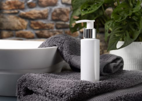 White cream squeeze tube on dark grey folded towels near vessel basin against stone wall and green plant in bathroom, close up, packaging mockup. Natural cosmetic product