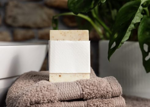 Soap bar with blank label on light brown folded towels, near green plant and vessel basin in bathroom, close up, brand packaging mockup. Men hygiene product