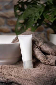 White cream squeeze tube on brown folded towels near vessel sink and green plant in bathroom, close up, packaging mockup. Natural cosmetic product for mens skin care