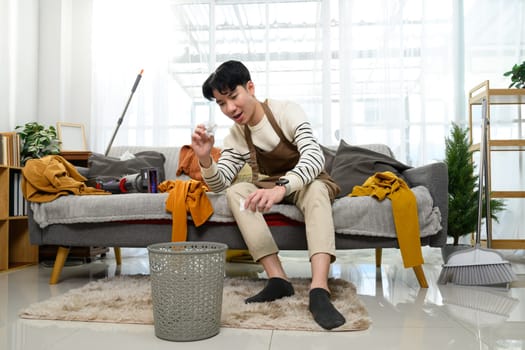 Man in an apron sitting in a cluttered living room and throwing crumpled paper into a trash.