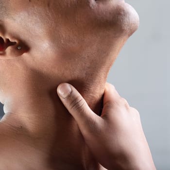 man grasps his throat with one hand, indicating possible throat pain or vocal cord discomfort. His head is slightly raised, and the close-up emphasizes the area of discomfort with a blurred background