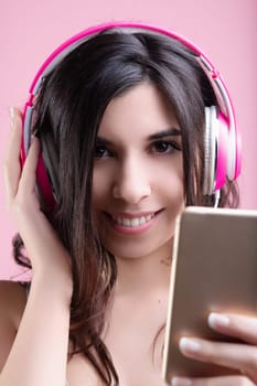 Young woman happily listens to music with pink headphones, holding a smartphone, looking relaxed and cheerful, enjoying wireless entertainment