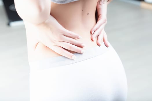 Woman's hands placed on her lower back, wearing casual white shorts and a grey top. This posture suggests she is experiencing discomfort or fatigue in the lumbar region
