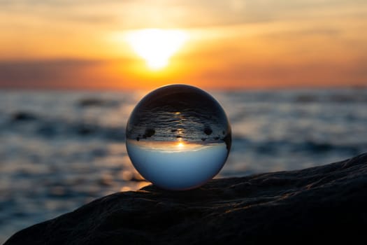Glass ball on a rock at sunset on the beach, the sea and the setting sun are reflected in the ball