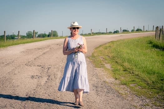 Happy mature woman is walking along a rural road on a bright summer day.