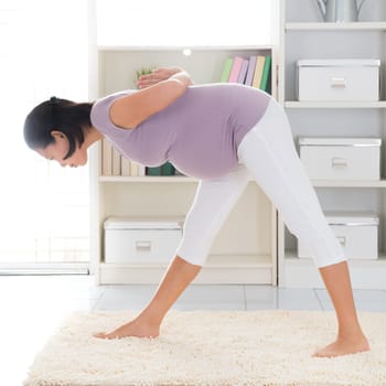 Prenatal yoga. Full length healthy 8 months pregnant calm Asian woman meditating or doing yoga exercise at home. Relaxation yoga forward bending pose.