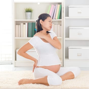 Shoulder or neck pain. Eight months pregnant Asian woman holding her back and shoulder while sitting on a floor at home.