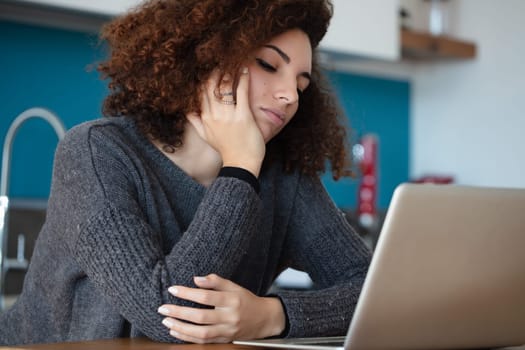 Woman with curly hair rests her face on her hand, staring at her laptop in frustration. The modern kitchen setting emphasizes her impatience, likely caused by buffering delays