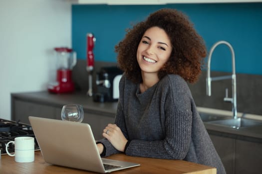 Young woman with curly hair is sitting at her kitchen counter, smiling while working on her laptop and enjoying a relaxing work from home experience