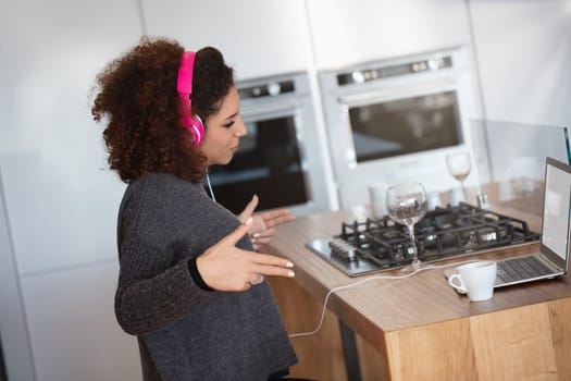 Woman with curly hair wears pink headphones in a modern kitchen, mimicking DJing motions. She sits by a laptop, with a cup and wine glasses nearby, creating a vibrant, club-like atmosphere