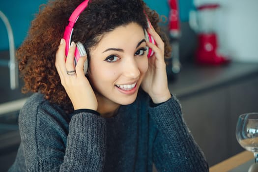 Young woman with curly hair enjoys music in her cozy kitchen, smiling and sipping a drink. The modern tech adds leisure to her home