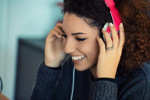 Happy young woman with curly hair enjoys music at home, smiling and immersed in the sounds with headphones. Her carefree expression captures modern relaxation and joy